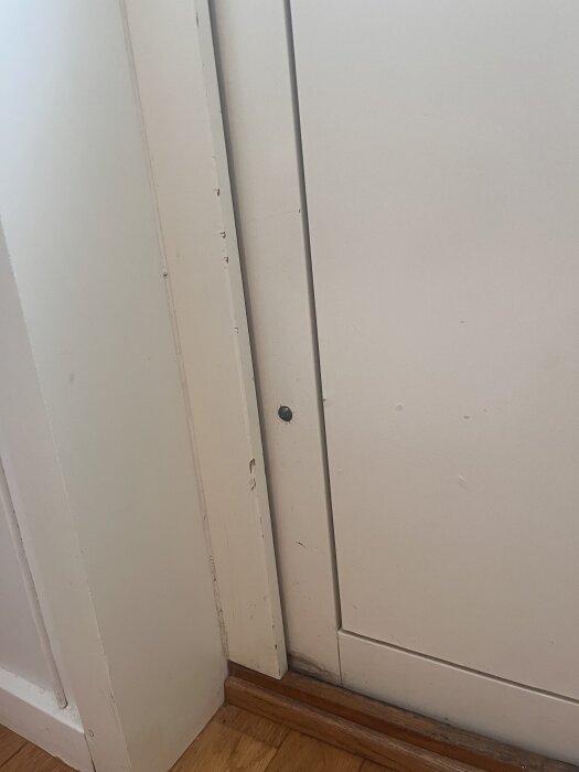 Närbild av vit innerdörr och dörrkarm med skador och skav i färgen, samt trälist vid golvets kant.