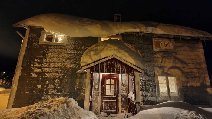 Hus täckt av tjockt snötäcke med snödrivor framför dörren och istappar hängande från taket i vinterlandskap.