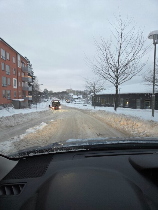 Bil på snöig väg i stadsmiljö, grå himmel, byggnader i bakgrunden och träd utan löv.