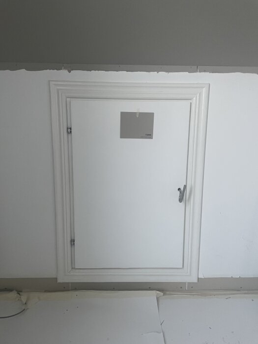 Vit dörr med list på snedvägg i sovrum, med färgprov fäst ovanför handtag. Renovationsprojekt med planer på akustikpanel och dörrmålning.