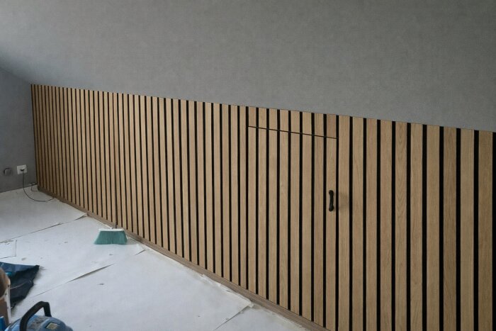 Akustikpanel på snedvägg med dörr till kattvind inkopplad, dörrhandtag synligt och konstruktion i ljust trä, i rum under renovering.