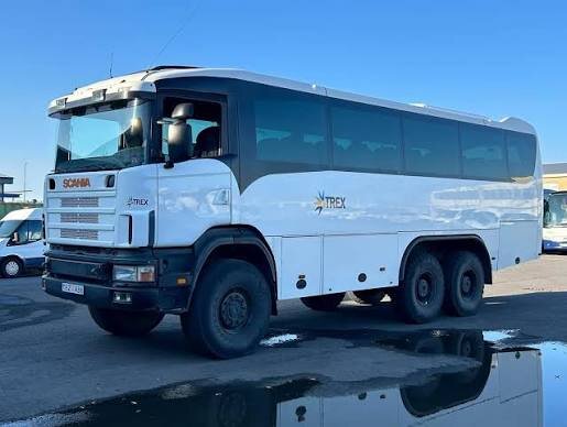 Vit fyrhjulsdriven buss från Scania parkerad på asfalt, reflekterad i en vattenpöl.
