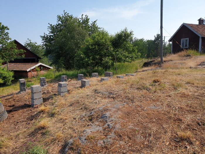 Byggplats med murblock utplacerade på marken, omgiven av träd och en ladugård i bakgrunden, under en klar himmel.