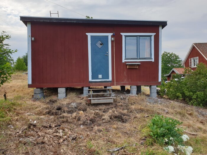 Röd bastubyggnad på plintar med blå dörr och fönster, omgiven av torrt gräs och buskar, stege och hus i bakgrunden.