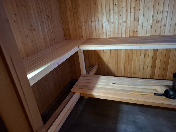 Vedeldad bastu med träpaneler och två lavbrädor, en skopa och slevar ligger på lavbrädan.