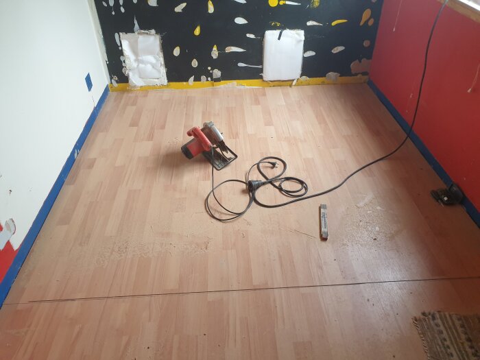 Rum under renovering med nylagt laminatgolv, röd och svart vägg med färgstänk, samt en eldriven handcirkel såg på golvet.