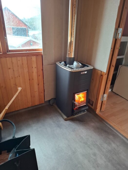 Vedeldad bastukamin i ett rum med träpanel och synligt fönster. En pågående eld syns genom kaminens glaslucka.