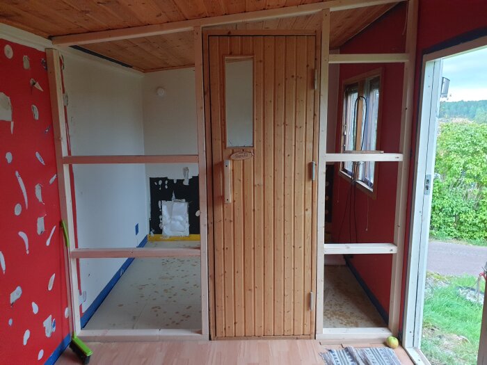 Vedeldad bastu under konstruktion med träpanel och delvis färdig inredning, öppet fönster i anslutande rum samt verktyg och material på golvet.