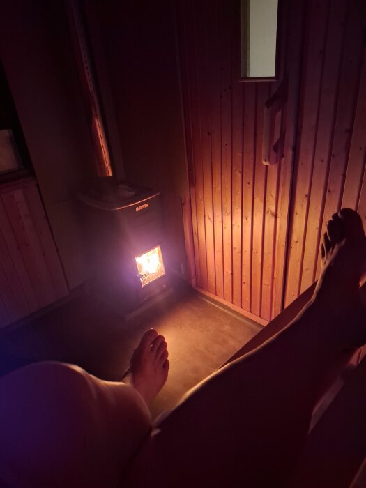 Person slappnar av i en vedeldad bastu med tända kaminen. Mörkt träinteriör, synliga fötter och varmt, dämpat ljus från elden.