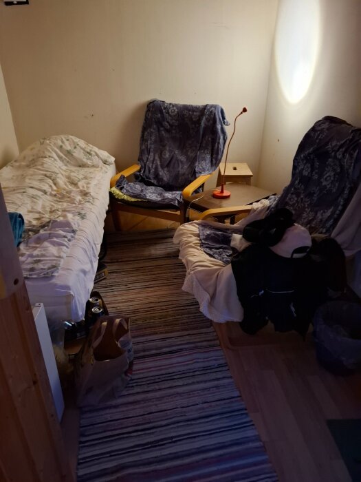 Ett rum med en säng, fåtöljer med filtar, ett litet bord med röd läslampa, randig matta och några papperskassar på golvet.