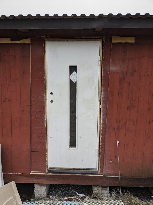 Röd vedeldad bastu med vit dörr och långsmalt fönster, ståendes på betongplattor, del av eget bastuprojekt.