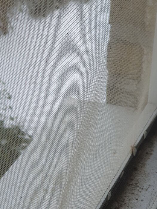 Fönsterbleck med löstagbara kanter bakom finmaskigt nät, del av husets fönsterdetalj, snö och murverk synligt genom nätet.
