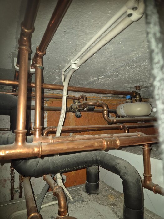 Rörsystem med kopparrör och isolerade rör i en källare, med en vit kabel och en ventil synlig.