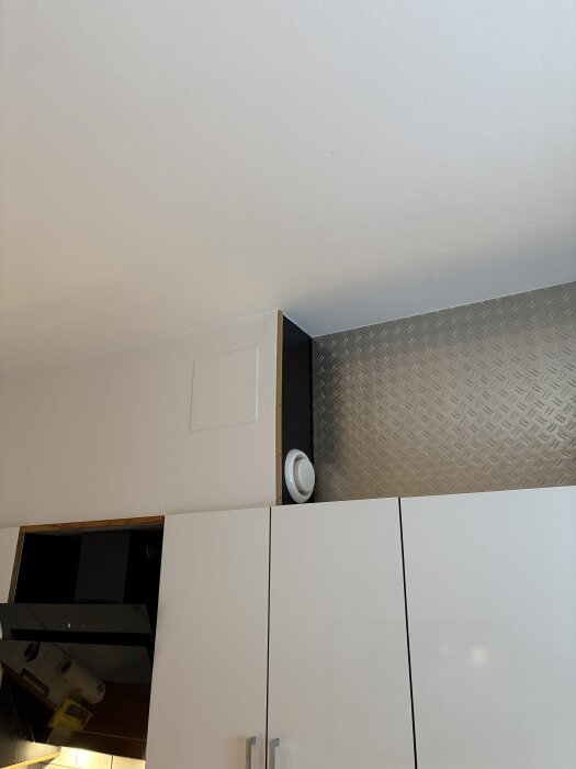 Köksvägg med vita skåp, ovanför syns ventilationsrör och ett litet ventilgaller. Taket är synligt ovanför skåpen.