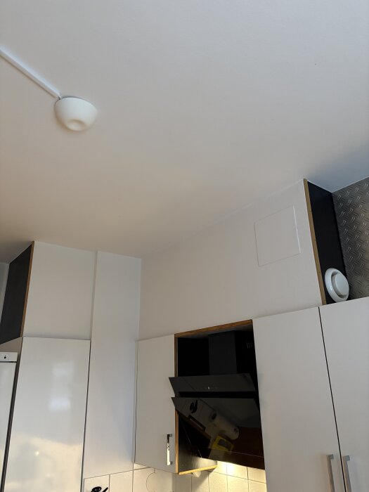 Kök med vit skåpinredning, synlig köksfläkt och överskåp nära taket. En vit ventilationsdetalj syns ovanför skåpen.
