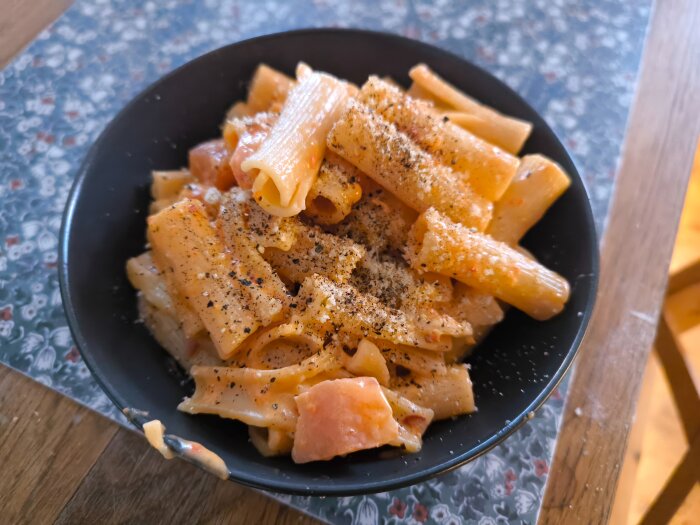 Rigatoni med kallrökt lax, Calabrese och ost toppad med svartpeppar i en svart tallrik.
