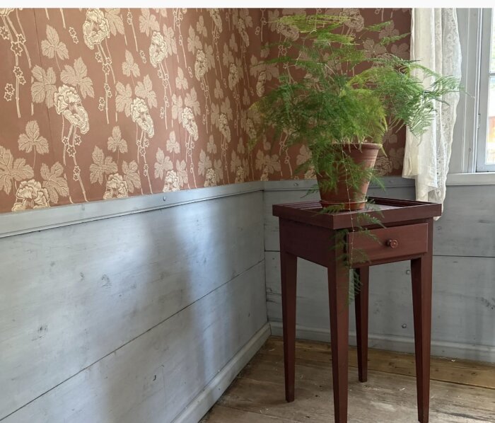 Vardagsrum med gammal slätpanel, blommig tapet och fönster. Ett mörkt träbord med grön växt står vid väggen.