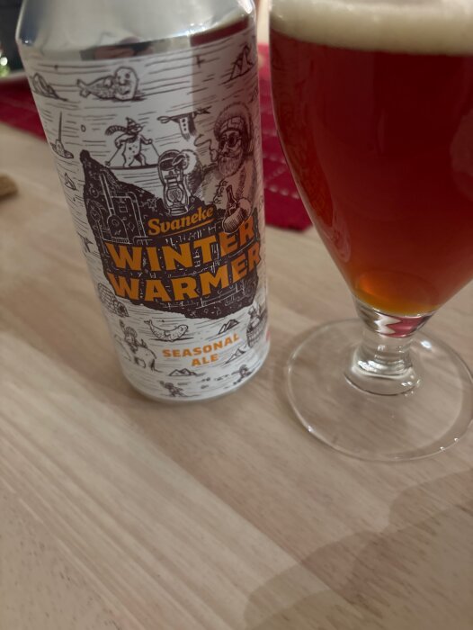 Burk med Svaneke Winter Warmer Seasonable Ale vid sidan av ett glas fyllt med mörk öl på ett bord.