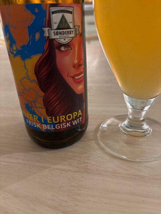 En flaska med etikett "Sønderby Bryghus" bredvid ett glas med ljus öl på ett ljust träbord.