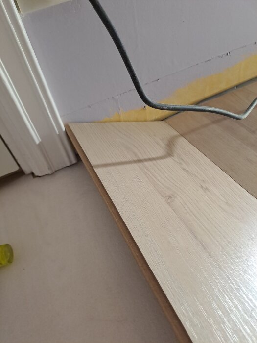 Ett ljust trälaminatgolv ligger delvis under en kapad dörrkarm. En krokformad verktygsliknande föremål syns också i bild.