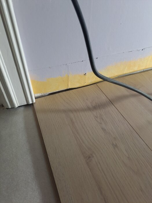 Golvläggning intill dörrkarm, bräda vänt mot dörröppning, del av karm borttagen nedertill för att få in brädan, svart kabel i förgrunden.