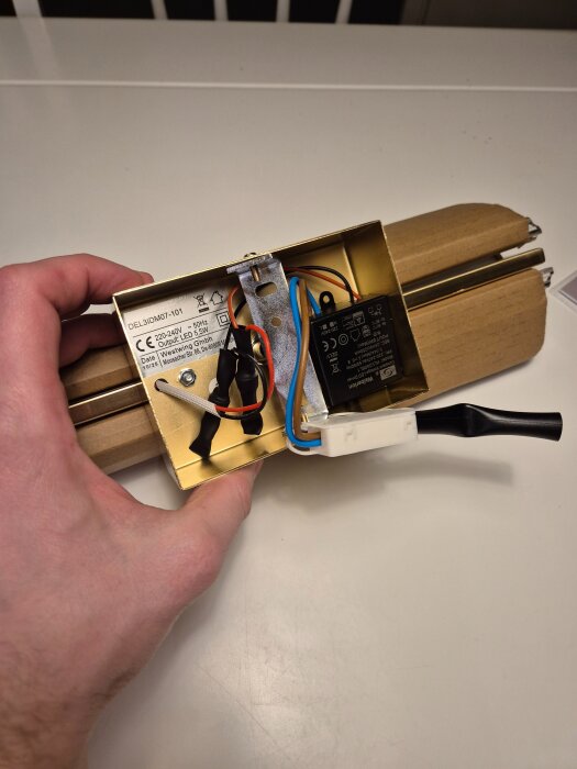 Närbild på baksidan av en vägglampa med synliga elektriska kopplingar och en hand som håller den guldfärgade metallhållaren.