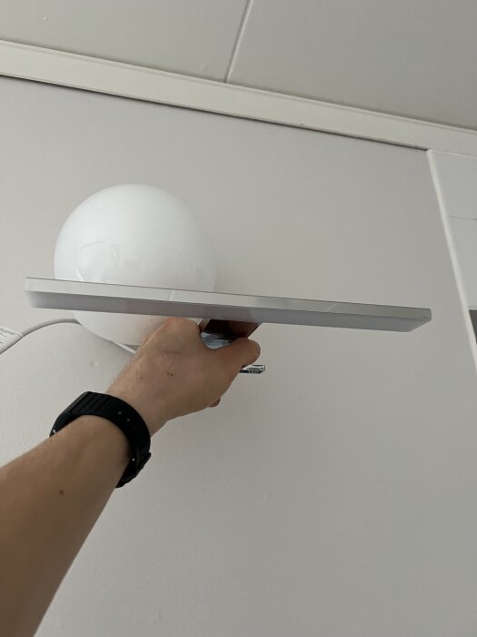 Hand som håller en lampa i tänkt placering ovanpå ett badrumsskåp mot en vit vägg, med tak och vägglist i bakgrunden.
