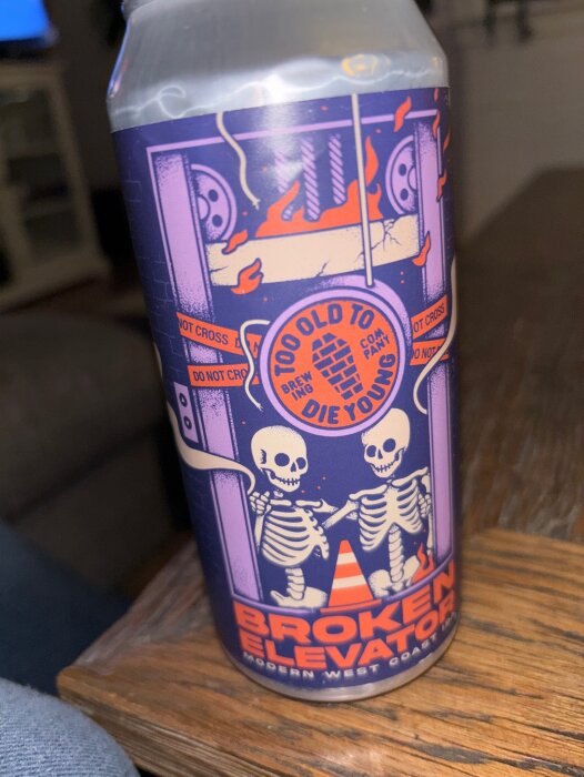 Burk med etikett för "Broken Elevator" IPA från Toooldtodieyoung, visar två skelett och eldlågor.