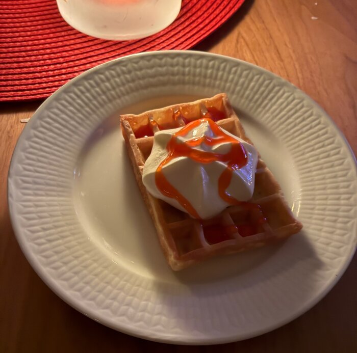 Våffla med grädde och sirap på en vit tallrik, ljusbrun träyta under och röd bordstablett i bakgrunden.