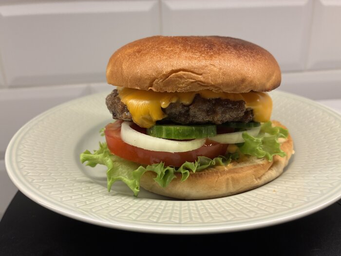 Hamburgare med ost, sallad, tomat, gurka och lök på en vit tallrik.