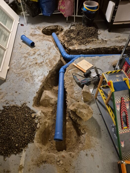 Blå plaströr installerade i marken under ett renoveringsprojekt i ett garage eller källare. Grus och verktyg syns runt omkring.