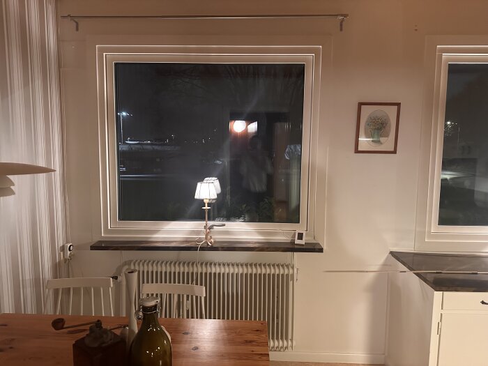 Gammal radiator under fönster i rum, med bord och stolar framför, lampor lyser i mörker.