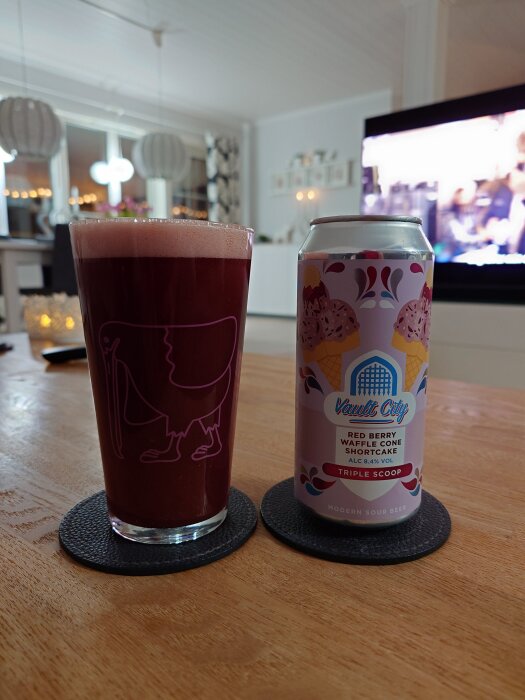 Ett glas mörkröd suröl och en burk med etiketten "Vault City Red Berry Waffle Cone Shortcake" på ett bord inomhus.