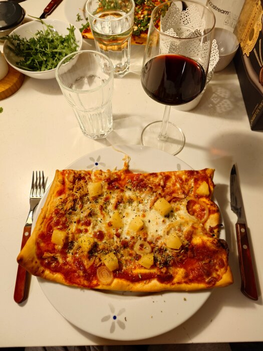 Pizzakit med tomatsås, ost, ananas och lök serverad på en vit tallrik, glas vin och sallad vid sidan.