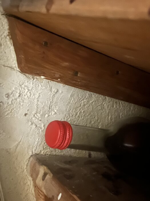 En röd kork på en flaska som ligger på en träyta, omgiven av fastspikade hyllor och en grov vägg.