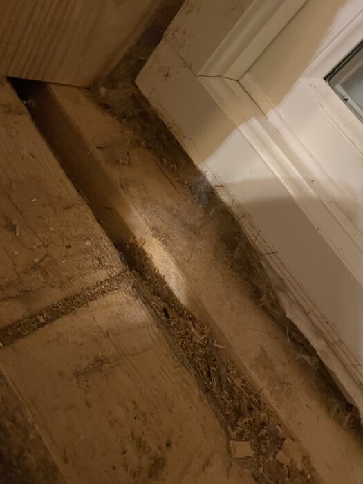 Spricka i trävägg vid fönsterkarmen, med spån och smuts som samlats runt öppningen.