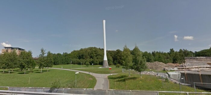 Vit, avlång skorsten på grön gräsmatta omgiven av träd och stigar under en blå himmel.