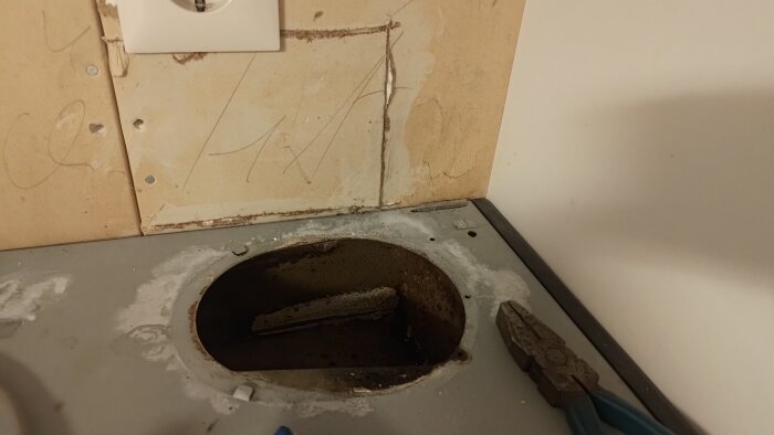 Gammalt köksuttag med trasig vägg, där en köksfläkt har som bortmonterats. En tång syns bredvid en öppning i bänkskivan.