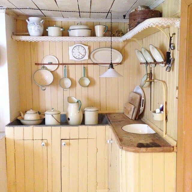 Ett gammaldags kök med beige panelvägg, porslinskärl, grytor, sil och väggklocka från tidigt 1900-tal.