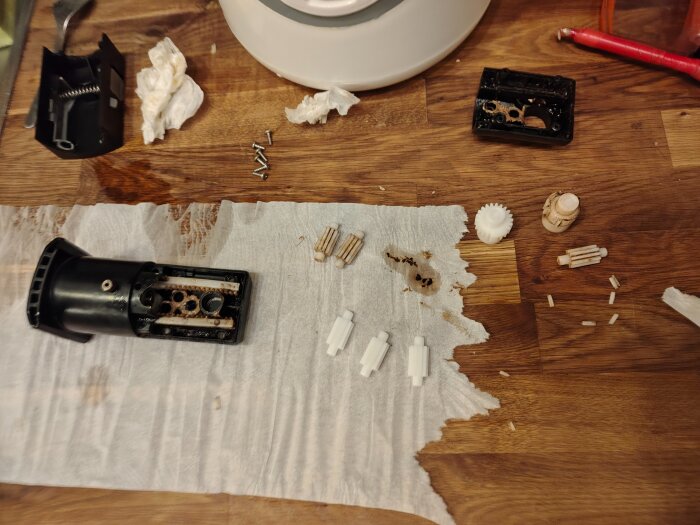 Delar från en isärplockad elektrisk apparat, inklusive drev och skruvar, ligger utspridda på ett köksbord med hushållspapper.