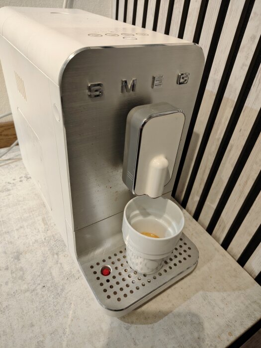 Vit espressomaskin med en kopp espresso under brygghuvudet, placerad på en köksbänk med randig bakgrund.