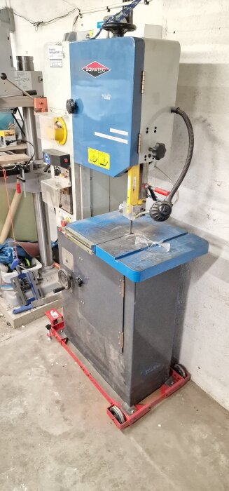 Bandsåg på en verkstadsvagn med blå och gul märkning. Skyddsglasögon ligger ovanpå sågen. Verktyg och utrustning syns i bakgrunden.