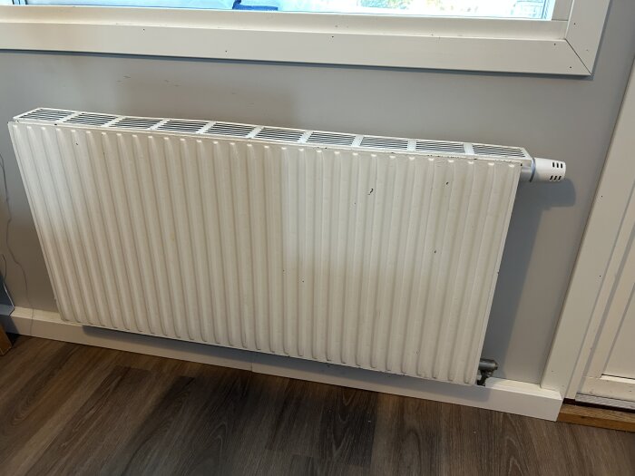 En vit radiator under ett fönster i ett rum med trägolv.