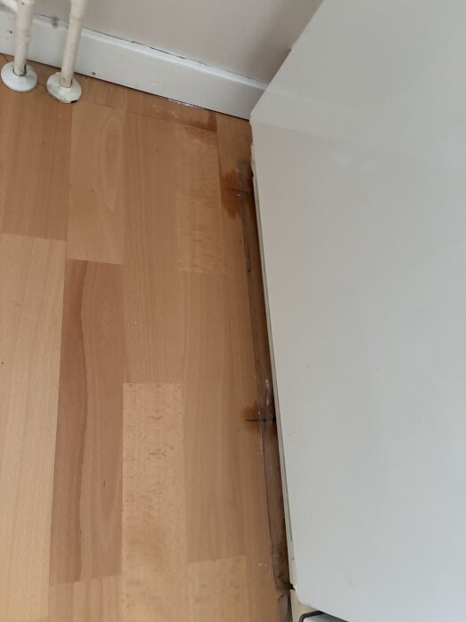 Missfärgad bokparkett med vattenskada vid ett hörn nära rör och vit vägg.