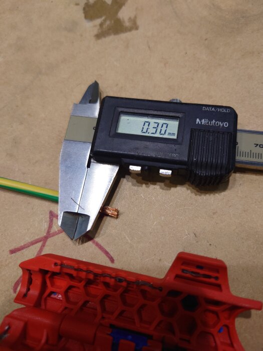Skjutmått mäter tjockleken av en kabel bredvid ett rött verktyg på ett bord, visar 0,30 mm.