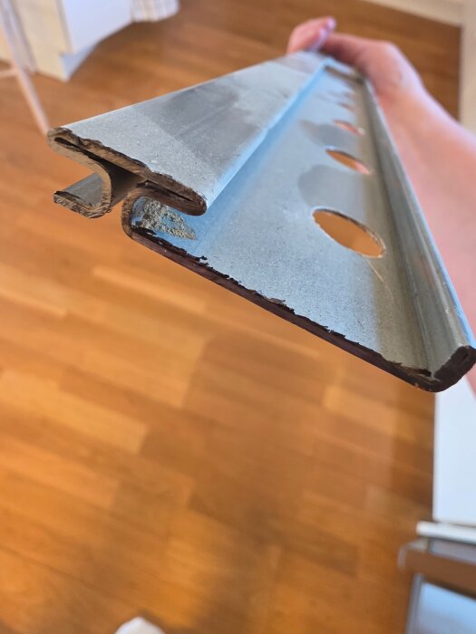 Närbild av en hand som håller en metod upphängningsskena, troligen för skåpmontering, med synliga skåror på båda sidor och flera runda hål.