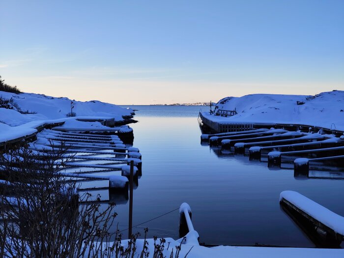 Snöklädd båthamn i Bohuslän, spegelklart vatten och vinterlandskap i skymning.