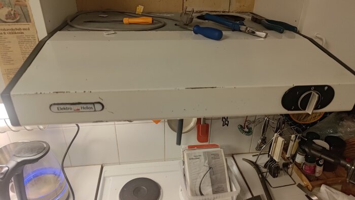 Fläkt av märket Elektro Helios monterad under köksskåp, med verktyg och sladdar synliga ovanpå, samt köksutrustning runtomkring.