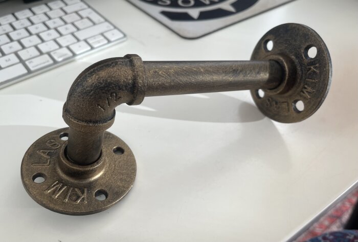 Metallkonsol i form av ett rör, skruvad ihop som hyllbärare. Industriell design, ligger på ett vitt bord framför ett tangentbord.