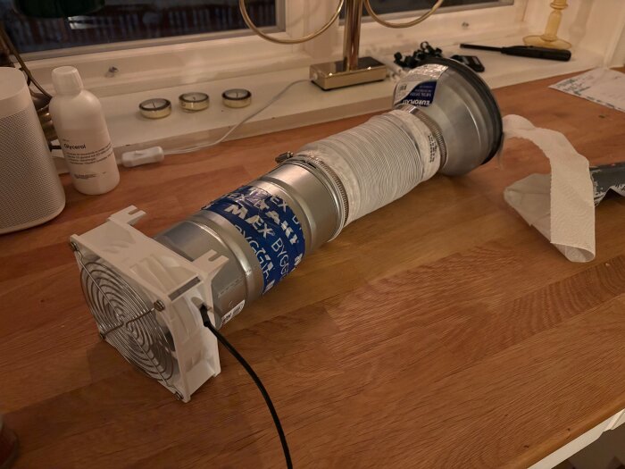 Hemmagjord ventilationslösning med fläkt och rördelar placerade på ett träbord, omgiven av diverse föremål som ljus och flaskor.